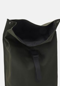 Sac étanche vert avec une texture douce, ouverture sur le dessus et doublure intérieure noire. Équipé d'une sangle noire et de boutons-pression pour la fermeture.