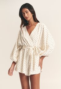 Robe blanche à pois noirs, avec un décolleté en V profond, de longues manches bouffantes et une taille nouée. Tissu léger et texturé.