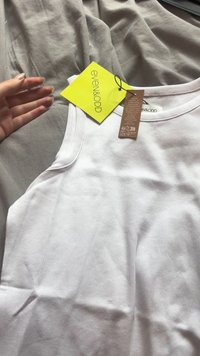 Weißes, geripptes Tanktop mit rundem Halsausschnitt und ärmellosem Design. Enthält ein gelbes Markenschild und ein braunes Pflegeetikett, das angebracht ist.