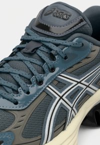 Gros plan d'une chaussure de course Asics bleue et grise montrant les lacets, la tige en mesh, les bandes du logo et la languette rembourrée.