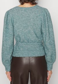 Turkos stickad tröja med strukturerad yta, ballongsärmar och midjebälte, kombinerad med bruna läderbyxor.