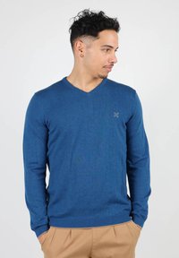 Pull bleu en V en tissu tricoté, avec des manches longues et des poignets côtelés. Un petit logo sur la poitrine ajoute un détail subtil.