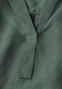 Primo piano di un tessuto in lino verde con un design a V, dotato di colletto piegato e texture morbida. Cuciture distintive visibili lungo i bordi.