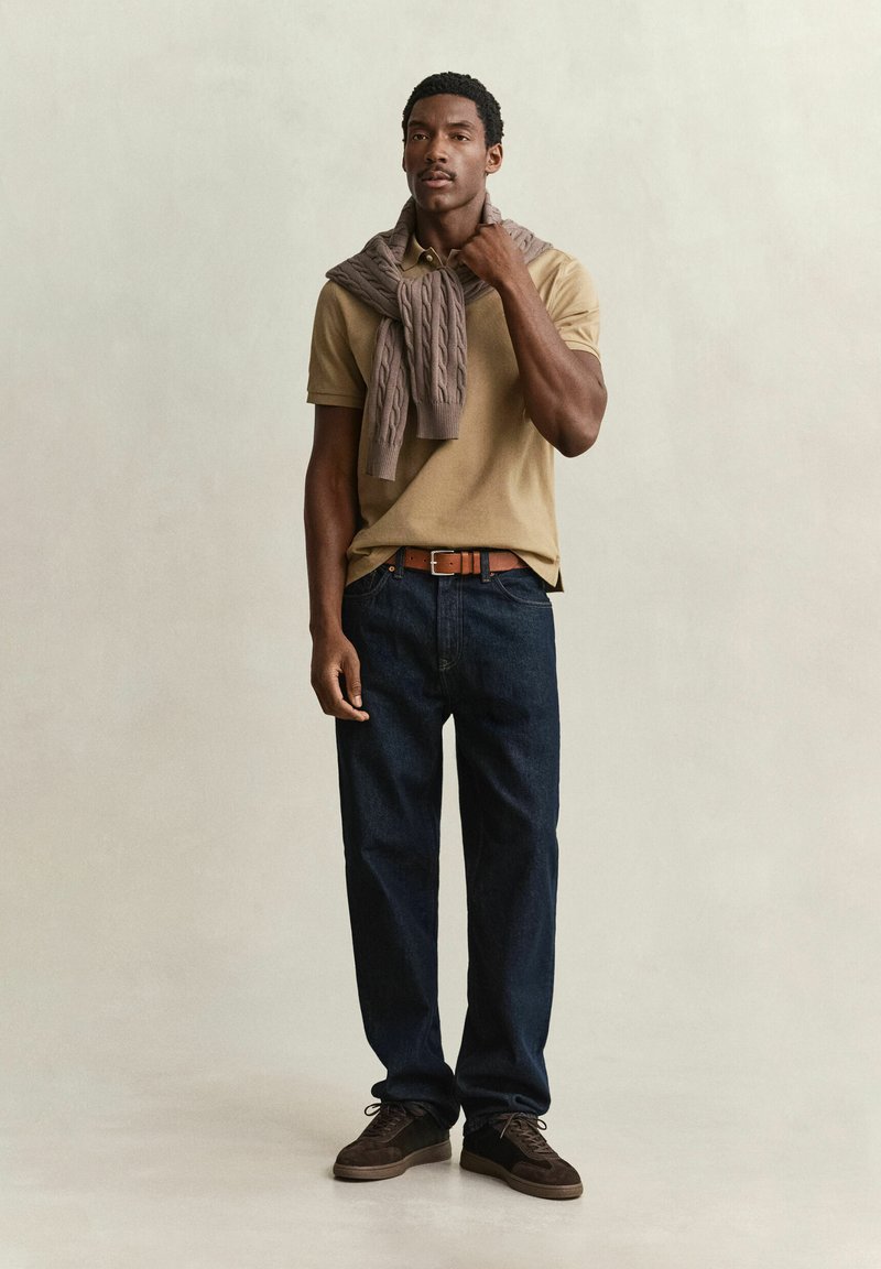 Homme portant un polo beige, un pull marron sur les épaules, un jean foncé, une ceinture marron et des chaussures marron, debout sur un fond uni.
