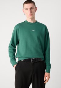Sudadera verde de manga larga con cuello acanalado, que presenta el logo blanco de "BOSS" en el pecho, combinada con pantalones negros y un cinturón negro.