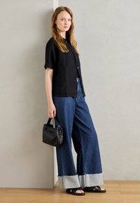 Svart kortärmad linneskjorta, blå vida jeans med fransar, svart vävd väska och svarta slidesandaler; minimalistisk design.
