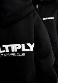 Černá mikina s kapucí, na které je v tučných bílých písmenech nápis "MULTIPLY" a menší text "FAMILY APPAREL CLUB", prezentující měkkou, texturovanou tkaninu.
