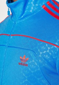 Modrá bunda na zip s červenými akcenty a třemi červenými pruhy na rameni. Na hrudi je vyšívané logo a materiál je texturovaný.