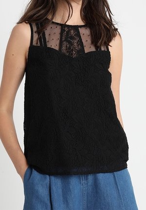 Kobieta w czarnym koronkowym topie bez rękawów z przezroczystym dekoltem w groszki i niebieskich spodniach z wysokim stanem, z rękami w kieszeniach.