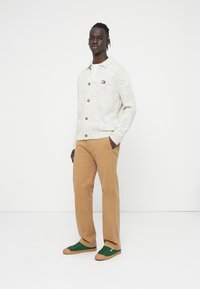 Μπεζ, πουά πλεκτή ζακέτα με ξύλινα κουμπιά, φορεμένη πάνω από λευκό πουκάμισο. Συνδυασμένη με καφέ παντελόνι και πράσινα αθλητικά παπούτσια.