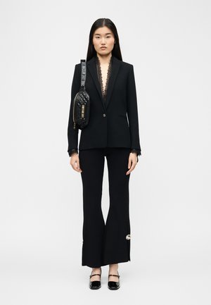 Kvinne iført svart skreddersydd blazer med blonderkant, svarte vide bukser med sideknapper, svarte Mary Jane-sko og en svart quiltet skulderveske.