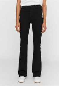Personne portant un jean bootcut noir avec des poches avant et des baskets blanches, debout devant un fond uni clair.