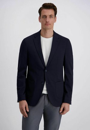 Man met kort bruin haar, een marineblauw colbert over een wit shirt en grijze broek, staande tegen een effen witte achtergrond.
