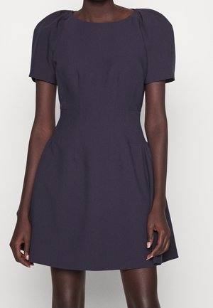 Kvinde iført en mørkeblå A-formet kjole med korte ærmer, puffede skuldre og en tætsiddende talje.