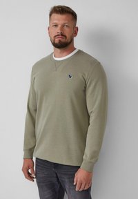 Felpa a maniche lunghe verde oliva realizzata in morbido cotone misto. Presenta un collo rotondo con un colletto interno bianco e una piccola toppa con logo.