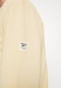 Ljusgul tröja med strukturerad stickning. Har en vit Reebok-etikett med texten "NATURAL DYE" på vänster ärm.