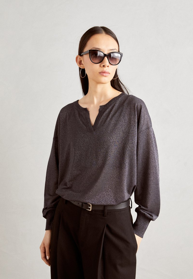 Locker sitzendes, dunkelgraues Oberteil mit einem subtilen Schimmer, das über einen V-Ausschnitt und lange Ärmel verfügt. Kombiniert mit schwarzen, hoch taillierten Hosen und Sonnenbrille.