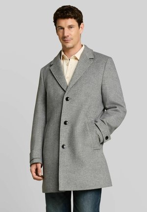 Uomo che indossa un cappotto grigio chiaro abbottonato con ampie falde, mano in tasca, sopra una camicia beige e jeans scuri, su sfondo neutro.