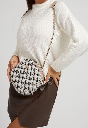 Femme portant un pull en maille blanc et une jupe en cuir marron, avec un sac bandoulière noir et blanc à motif, à bandoulière chaîne dorée.