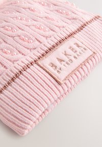 Strikket lyserød hue med ribbet tekstur, hævede mønstre og dekorative perle-lignende detaljer. Inkluderer et beige mærke med "BAKER" skrevet med gyldne bogstaver.