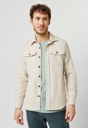 Camicia button-up color beige chiaro con due tasche sul petto, bottoni marroni e colletto classico. Il tessuto sembra morbido con una finitura testurizzata.