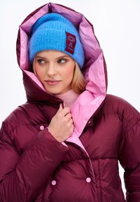Burgundowa kurtka puchowa z różowym podbiciem, z akcentami w postaci guzików. Jasnoniebieska czapka z czerwonym metką z napisem "ZOBACZ WSZYSTKO". Wzór z teksturowanego dzianiny.