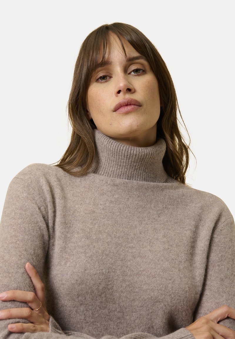 Maglione a collo alto color marrone chiaro con colletto a coste, texture morbida e vestibilità rilassata. Braccia incrociate, dettagli minori vicino ai polsini.