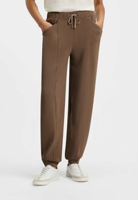 Bruine, elastische broek met taillekoord, voorzien van zijzakken en taps toelopende manchetten. Gladde textuur met contrasterende verticale naden.