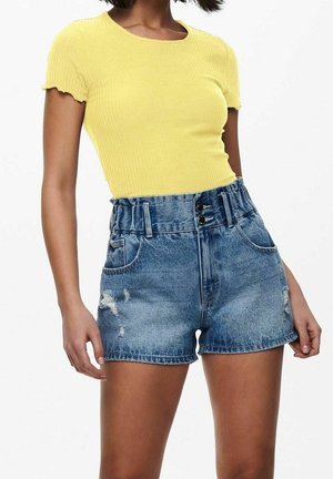 Kobieta ubrana w żółty prążkowany top z krótkim rękawem oraz wysokiej talii niebieskie szorty jeansowe z falbanką w pasie i postrzępionymi detalami.