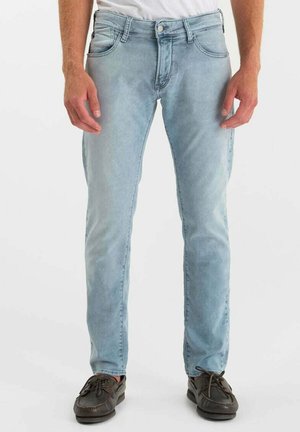 Man draagt lichtblauwe slim-fit jeans en donkerbruine veterschoenen, staand tegen een effen witte achtergrond.