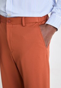 Pantalons orange texturés au fit décontracté, avec poches latérales et une taille avec passants de ceinture ; associés à une chemise rayée bleu clair.