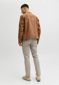 Brązowa zamszowa kurtka o klasycznym kroju, beżowe spodnie i jasne buty. Tło jest jednolite i neutralne.