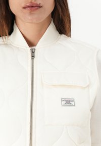 Gilet imbottito color crema con colletto alto, chiusura a zip e taschino sul petto. Presenta un'etichetta con logo e dettagli in tessuto testurizzato.