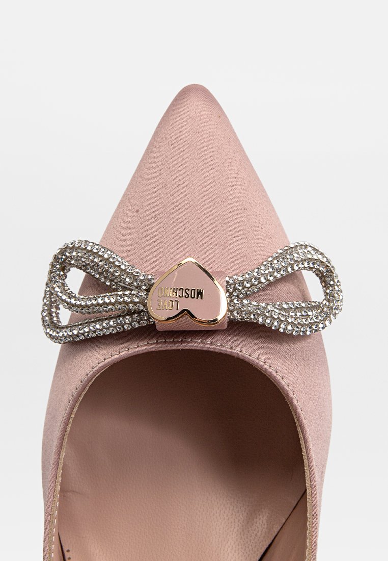 Décolleté in satin rosa-beige con punta affusolata, caratterizzato da un fiocco decorato con strass e un dettaglio a forma di cuore dorato con il nome del marchio.