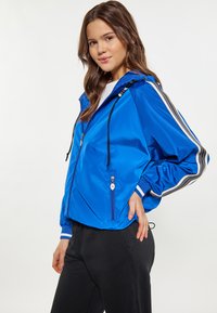 Giacca a vento blu con cappuccio, strisce atletiche nere e bianche, tasche frontali con cerniera e polsini elastici. Materiale liscio e leggero.