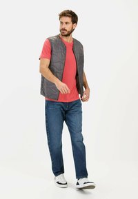 Grå quiltet vest, rød T-shirt, blå jeans og hvide sneakers. Vesten har en frontlynlås og en afslappet pasform.