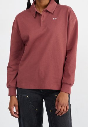 Persoon draagt een long-sleeve Nike polo in stofroze en zwarte spijkerbroek met details op de voorkant, staand tegen een effen achtergrond.