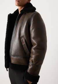 Giacca in pelle marrone con colletto in shearling nero, chiusura laterale con zip e finitura testurizzata; presenta dettagli in pelle nera e tasca a toppa.