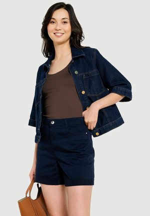 Usmievavá žena v tmavom riflej sakom cez hnedé tielko a tmavomodré kraťasy, drží béžovú kabelku na jednotnom svetlom pozadí.