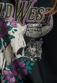 Tissu noir avec un design graphique comprenant "WILD WEST" en vert, des fleurs violettes et un paysage désertique avec un croissant de lune.