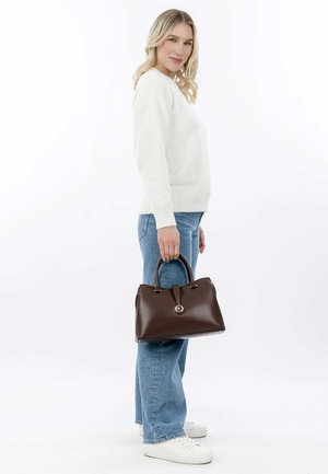 Borsa in pelle marrone con forma strutturata e manici arrotondati, dotata di un accento metallico centrale. Indossata da una persona in abbigliamento casual.