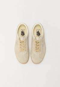 Beige suedesneakers med et klassisk design, som har lysebrune snørebånd og en såle i gummigummi. Detaljene inkluderer broderede elementer og en logomerke.