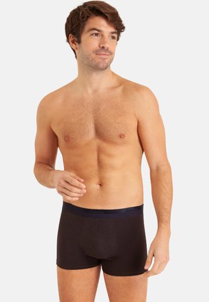 Homme torse nu à la peau claire et aux cheveux bruns, portant un boxer noir, debout et regardant légèrement sur le côté sur un fond uni.