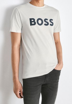 Persoon draagt een lichtbeige T-shirt met het woord "BOSS" in grote zwarte letters op de borst, gecombineerd met donkergrijze broek.