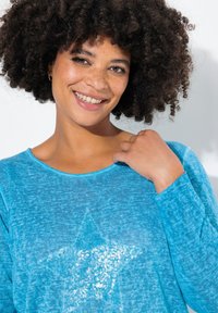 Top a maniche lunghe turchese con un design a stella in paillettes. Tessuto morbido e strutturato con scollo rotondo e vestibilità rilassata.