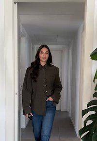Oversized, mørkebrunt rutete skjorte med knapper og lange ermer, kombinert med blå jeans. Innendørs miljø med en plante i nærheten.
