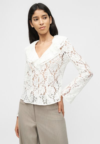 Γυναίκα που φορά λευκό μπλουζάκι με  λουλουδάτο δαντελωτό σχέδιο και γιακά με βολάν, μαζί με μπεζ παντελόνι με λεπτές ρίγες, ποζάροντας με το ένα χέρι στη μέση.