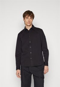 Camisa de manga larga negra con un frente de botonadura, que presenta un cuello clásico y una tela texturizada. Los puños están ajustados y adaptados.