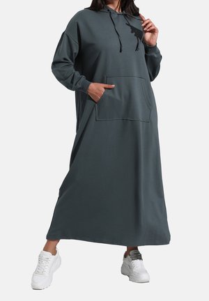 Osoba ubrana w ciemnoszarą, długą sukienkę z kapturem i przednią kieszenią oraz białe sneakersy, trzymająca za jedną sznurówkę kaptura, a drugą rękę w kieszeni.
