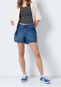 Džínové šortky tmavě modré barvy s vysokým pasem a bočním kapsou; nošeny s šedým upnutým topem a krémovým oversized sáčkem. Bílé tenisky dotvářejí celý outfit.